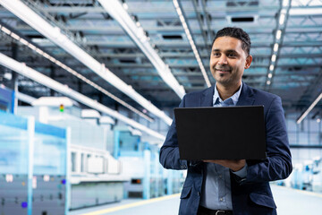 Fototapeta premium Smiling Indian man working in microchip factory assessing hardware performance using laptop. Happy semiconductors manufacturing plant manager optimizing supply chain logistics with notebook
