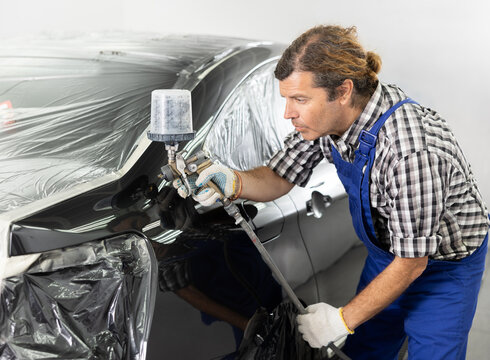 Experienced male auto painter in blue overalls working in car repair workshop, focused on painting covered with film black vehicle using spray gun..