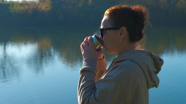 Woman drinking hot coffee from thermos cup by a serene lake. Profile of a relaxed woman in a hoodie and sunglasses enjoying a warm drink from a thermos cup while standing by a tranquil lake