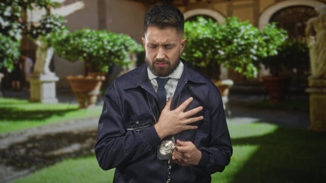 Man detective wearing badge and holding handcuffs clutching hand to chest in a building courtyard with statues and planters; duty concern.