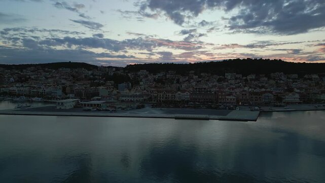 A coastal town at dusk, with boats in the harbor and buildings nestled against a tree-covered hill under a colorful sky.