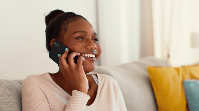 An African American woman enjoys a conversation on her phone while seated on a cozy couch. The room is well-lit with colorful pillows in the background, creating a warm atmosphere.