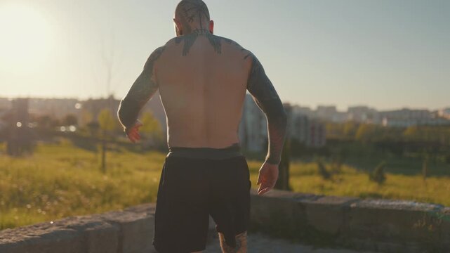 Tattooed muscular man walking away at sunset