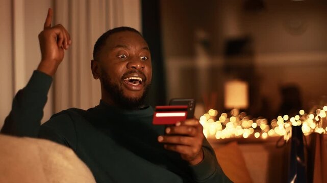 A man with dark skin sits on a couch indoors, holding credit cards while using his smartphone. Warm lights and a cozy atmosphere fill the background. He looks thoughtful as he focuses on his device.