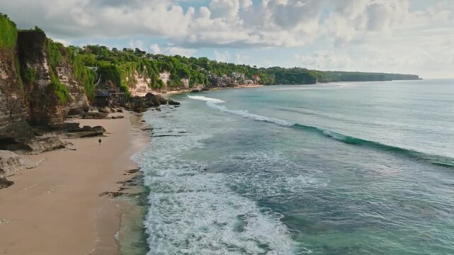 Indonesian coastline aerial view with sandy beach and lush green cliffs meeting turquoise ocean, lonly person walking along the shore as waves break over the shallow reef. Aerial view drone flight