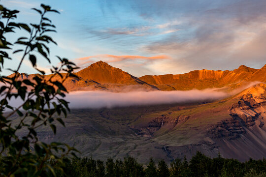 Wisps of low fog drift lazily across the valley as the early morning sun casts a warm golden glow onto the rugged volcanic mountain ridges of Skaftafell in Southern Iceland