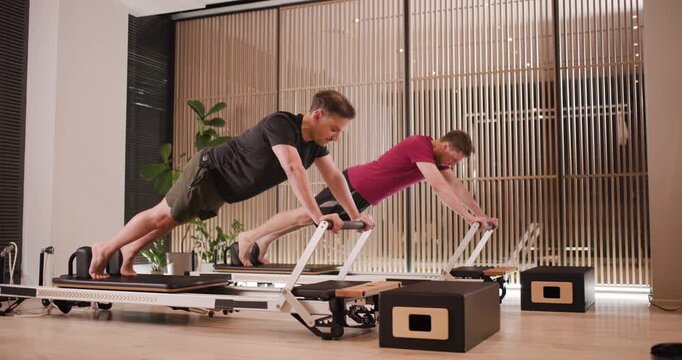 Two individuals engage in a vigorous workout using reformer machines in a stylish studio. The environment features natural light and greenery, enhancing the fitness experience
