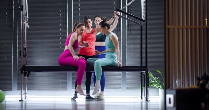 Three women engage in a fitness discussion while sitting on equipment in a bright and modern studio. They are dressed in colorful workout attire, sharing tips and encouragement