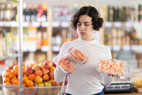 Woman buyer doubts and hesitates, chooses and select prawn in supermarket. Before purchasing, evaluates product, selects appropriate item, check expiry date, look out seafood delicious.