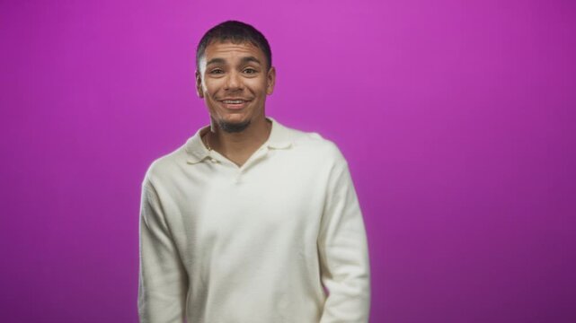 Man touching chest with hand, wearing white sweater and making expressive gestures on a purple studio backdrop; uncertainty introspection.