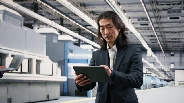 Portrait of cheerful senior microchip factory manager using tablet to conduct study on components. Jolly Asian executive walks in PCB production plant, overseeing semiconductors with device, camera B