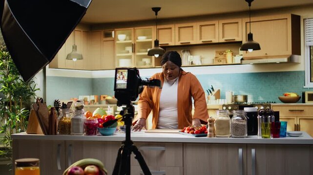 Behind the scenes of black woman recording meal prep vlog with tripod in the kitchen, presenting cooking process for online social media audience. Female influencer and content creator.