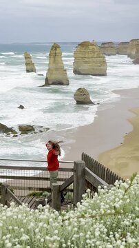 Traveler girl stands at the viewpoint spreading her hands and admiring powerful waves crashing against the limestone stacks of the Twelve Apostles on the Great Ocean Road, Victoria, Australia