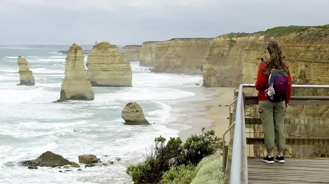Traveler girl stands at the viewpoint and admires powerful waves crashing against the limestone stacks of the Twelve Apostles on the Great Ocean Road, Victoria, Australia