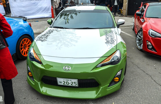 Green and white Scion FR-S sports coupe with unique hood graphics, lowered suspension, custom aerodynamics, and yellow fog lights displayed among other auto show exhibits