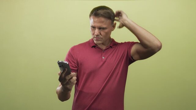 Man holding smartphone and scratching head in studio while wearing red polo, looking at screen; confusion doubt.
