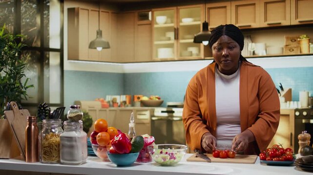 African american woman chopping cherry tomatoes on cutting board and adding ingredients in a bowl to make fresh salad for online food vlog tutorial. Influencer making culinary content.