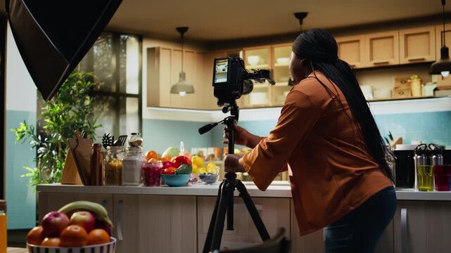 Food content creator fine tuning camera settings before filming recipe video, behind the scenes footage with black woman preparing studio lighting and tripod placement for smooth food vlog.