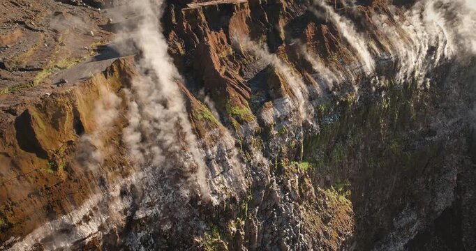 Ambrym Island volcano crater rim with steaming fumaroles, rugged layered lava walls, green moss patches and white gas plumes rising from vents under bright sunlight. Travel to Vanuatu. Aerial shot
