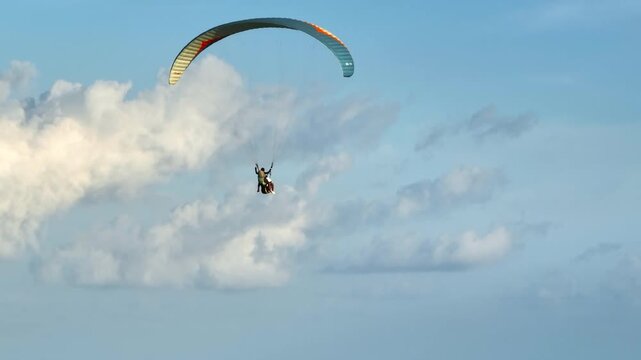 Two people enjoying a tandem paragliding flight against a clear blue sky with white clouds, experiencing freedom and adventure.