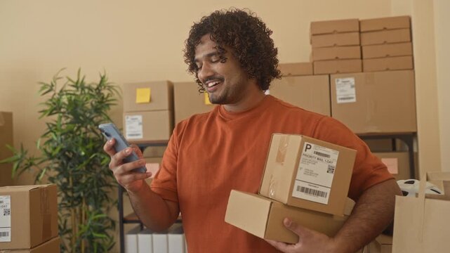 Man holds smartphone and cardboard packages, smiling and checking labels while standing in a building; confidence.