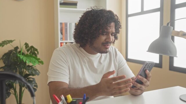 Man tapping smartphone screen and gesturing with raised hand at desk in building; frustration tech issue.