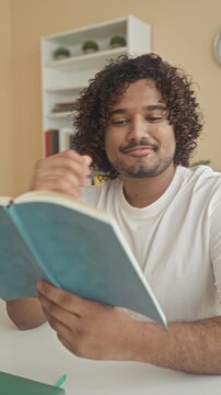 Young indian man reading book at a library table, hand turning page and hand to chin gesture by bookshelf; thoughtful focus.