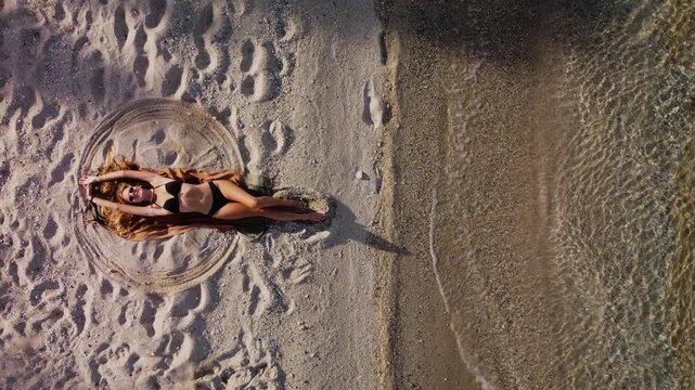 Woman in Bikini Making Snow Angel on Sandy Beach by the Ocean, Greece