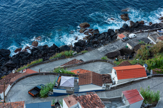 Road to Farol do Arnel. Winding coastal road descends past red-tiled houses. Waves crash on the rocky shoreline. High-angle view of ocean and cliffs. Sao Miguel. Azores. Portugal.