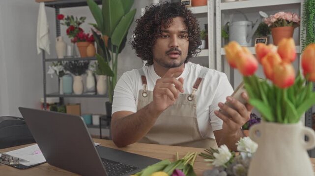 Man florist in apron holds smartphone and taps screen while checking laptop and arranging flowers in studio; small business concentration.