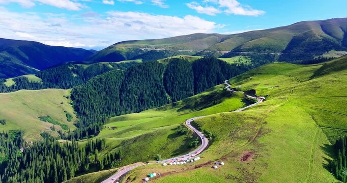 Aerial view of a winding asphalt road through lush green forests and scenic alpine hills in Xinjiang, China.