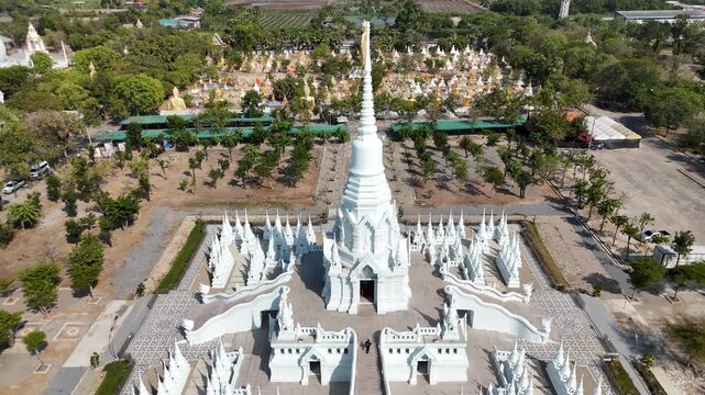 A cinematic drone shot featuring a smooth diagonal descent towards the stunningly symmetrical white Chedi Roi Yot pagoda, also known as the Temple of a Hundred Spires, in Suphan Buri, Thailand.