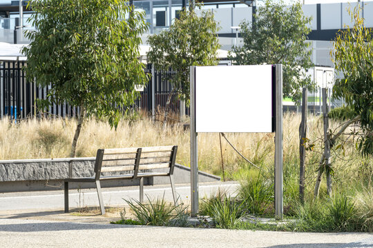 Blank signboard or empty white mockup notice board beside a bench in a landscaped public space in Australia, with plants around footpath. Background texture of wayfinding, public amenities information