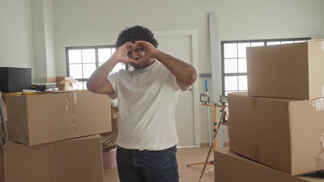 Man in white tshirt makes heart gesture with hands amid stacked moving boxes in building interior while smiling and unpacking; love fresh start.