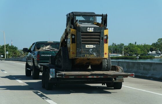 Bradenton, Florida, U.S.A - March 27, 2026 - Green Dodge pickup truck towing a yellow CAT skid steer on a flatbed trailer along a highway bridge.