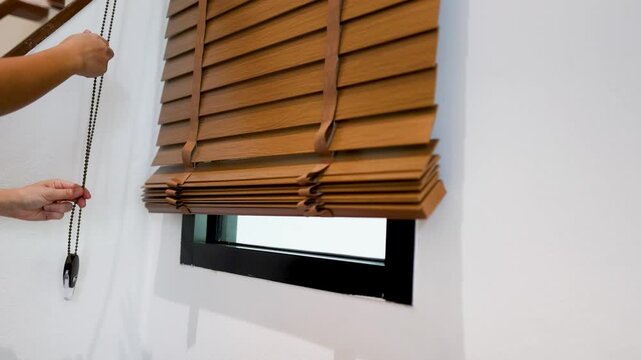 Hands Pulling Cord to Raise Wooden Venetian Blinds on White Wall