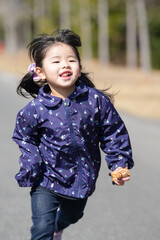 Fototapeta premium Happy child runs joyfully through sunlit park path. Her laughter echoes as wind tugs at her dark hair. Purple jacket adorned with star patterns adds charm. Her smile radiates pure, unguarded delight
