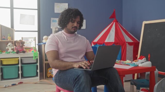 Man teacher seated at small chair typing on laptop, fingers on keyboard in kindergarten building; serenity focus.