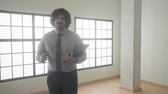 Man holding black folder and gesturing with hand in an empty building room while presenting property for sale; confidence.