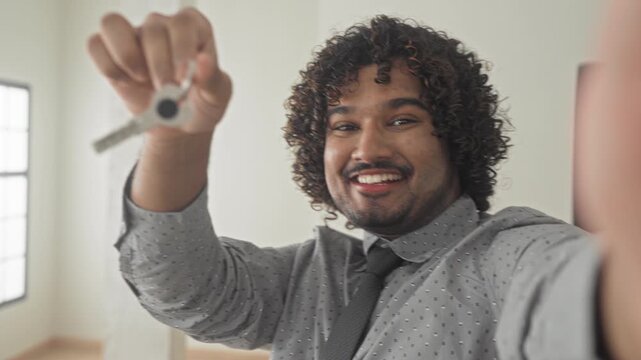 Man in shirt and tie holds key up with hand toward camera in building, smiling while offering house key; homeownership joy.