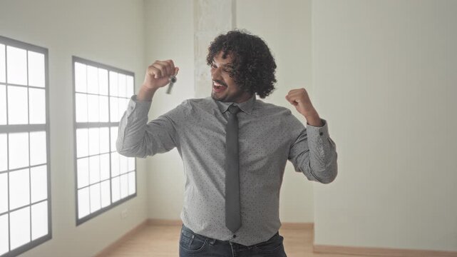 Man holding house keys in hand, smiling and raising fist beside tall windows in empty building room with wooden floor; homeownership pride.