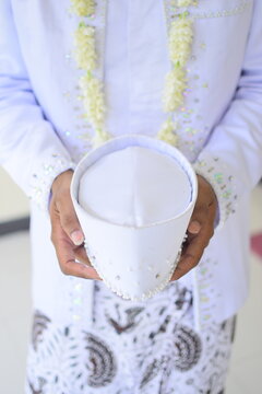Close-up of an Indonesian groom holding a white wedding peci hat with traditional jasmine garland.