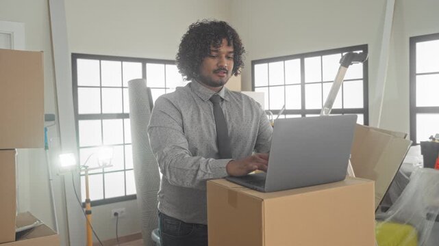 Man typing on laptop with hands on keyboard in building surrounded by moving boxes and a ladder; new setup focus.