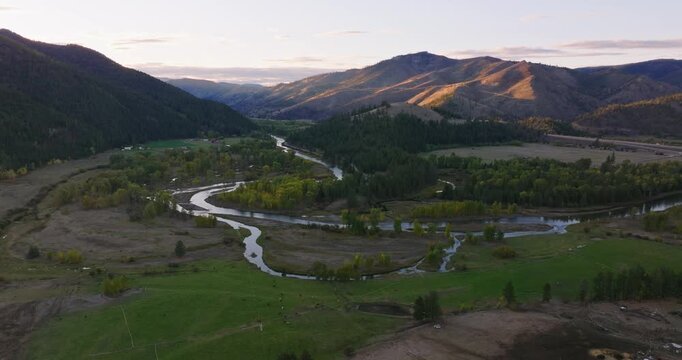 Winding river crossing wide rural valley with forest patches and farmland beneath soft evening sky, aerial drone, ascend over park