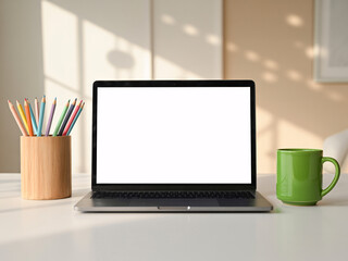 Fototapeta premium A modern workspace with a laptop, colorful pencils, and a green coffee mug on a clean white desk in a bright room.