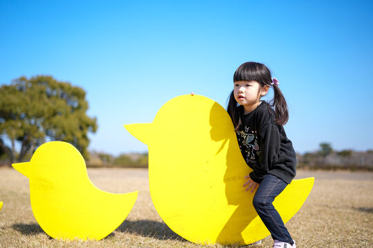 Young girl leans against bright yellow duck-shaped cutouts. Her pigtails bounce as she smiles at the camera. Clear blue sky frames the open grassy field. Sunny day enhances cheerful, playful mood
