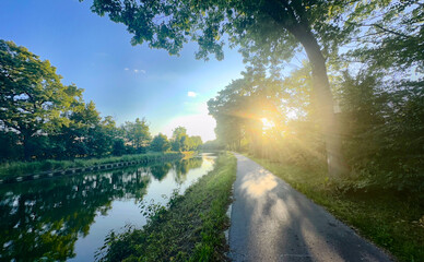 Rijkevorsel, Antwerpse Kempen, Belgium, Sunburst over canal path with golden rays cutting through trees, shimmering water reflections, warm glow on walkway and lush © Bjorn B