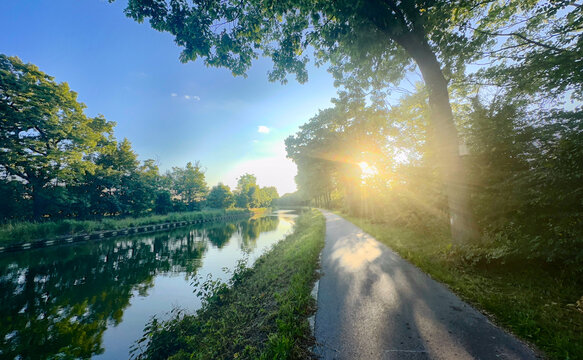 Rijkevorsel, Antwerpse Kempen, Belgium, Sunburst over canal path with golden rays cutting through trees, shimmering water reflections, warm glow on walkway and lush