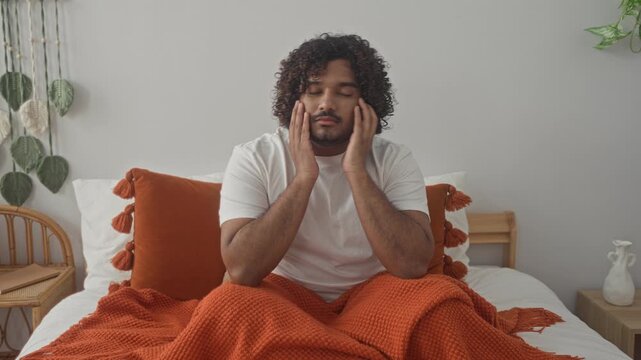 Man seated on bed touching his cheeks with both hands, wearing a white tshirt and wrapped in an orange blanket in building; tired reflection.