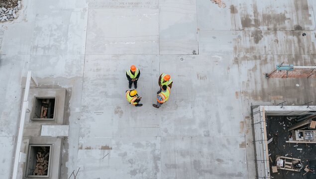Aerial perspective of four construction workers in safety gear collaborating on a vast concrete site, engaged in a strategic discussion about project development and on-site management